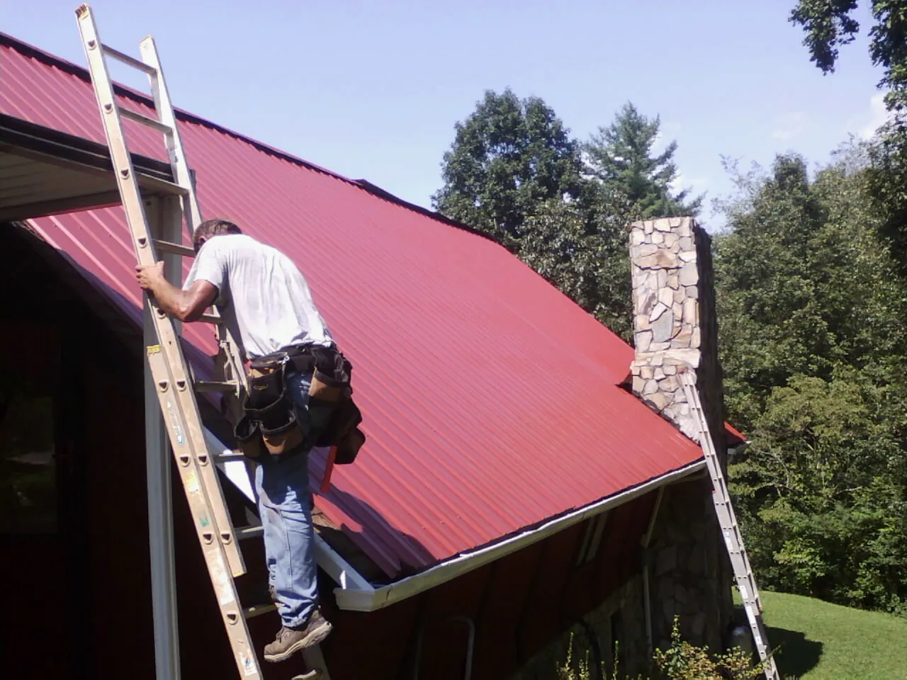 Roofer on a ladder beside a red metal roof and stone chimney — Soffit & Fascia Repair by Roofing Brothers in Trappe